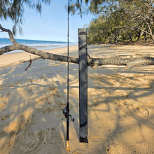 Load image into Gallery viewer, A black and gray vertical fishing rod holder standing on a sandy beach, holding a fishing rod upright. 